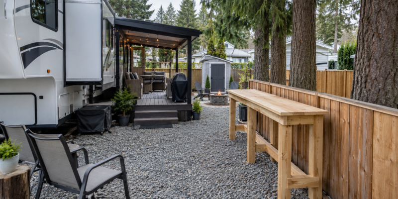 Unfinished DIY wooden outdoor bar beside a fence on a small RV patio lot with a fifth wheel, covered seating area and large trees.